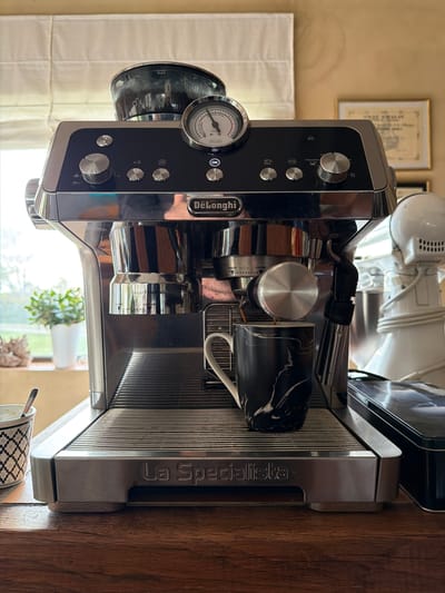 An indoor scene, presumably a kitchen in Mauléon-d'Armagnac, France, depicts a brushed stainless steel and black DeLonghi La Specialista espresso machine centered on a dark brown wooden countertop. The machine is actively dispensing a dark liquid, presumably coffee, from its portafilter into a black and white marbled ceramic mug positioned on the drip tray. The machine's control panel features a prominent pressure gauge with a needle indicating a setting within the "Optimal Zone," several silver metallic buttons including an "OK" button, a power icon, "x2" for double shot, "Espresso," and "MY" for custom settings, and two circular metallic knobs. Above the control panel, a transparent plastic bean hopper with a visible "MAX" fill line is partially visible. Reflections on the machine's polished surfaces show indistinct elements of the room and possibly a person.
To the left of the espresso machine, a white window with horizontal white Roman blinds allows ambient light into the room. Outside the window, green foliage and a white potted plant are visible. A small white and black geometrically patterned cup containing a spoon is positioned on the countertop to the far left. To the right of the machine, a white stand mixer with coiled cords is partially visible against a light beige wall. A gold-framed document or certificate hangs on the wall. A rectangular black object, possibly a storage tin or base for another appliance, sits on the countertop next to the mixer. The wooden countertop displays a distinct grain pattern.