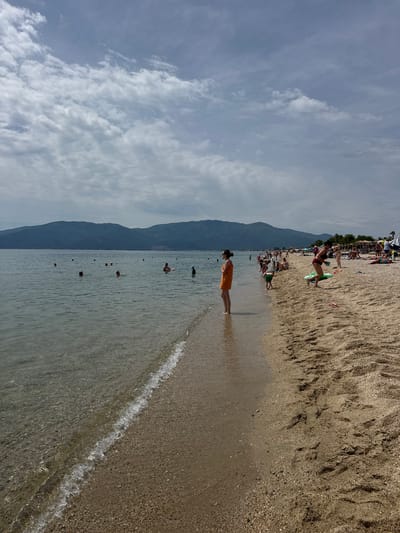 This is a wide shot of a beach in Asprovalta, Greece, on a cloudy day. The foreground shows the shoreline with clear water lapping onto the sand, which is covered in small pebbles. In the mid-ground, several people are in the water, swimming or wading. Further back on the sandy beach, more people are scattered, some sitting under umbrellas, others sunbathing or standing. In the far background, a range of green mountains rises against the cloudy sky. A single woman in an orange dress stands at the edge of the water, looking out towards the sea. The overall atmosphere is one of a leisurely summer day at the beach.