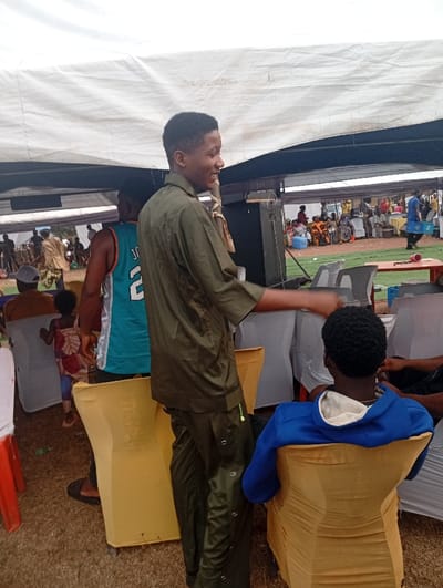 An outdoor event shows a smiling young man in an olive-green outfit standing and gesturing. Behind him, another man in a basketball jersey stands next to a child. A person sits in a yellow-covered chair to the right, with a blue hoodie visible. In the background, under a large white tent, many people are seated, suggesting a gathering. Chairs are arranged in rows, and a speaker system is visible. The ground appears to be grassy.