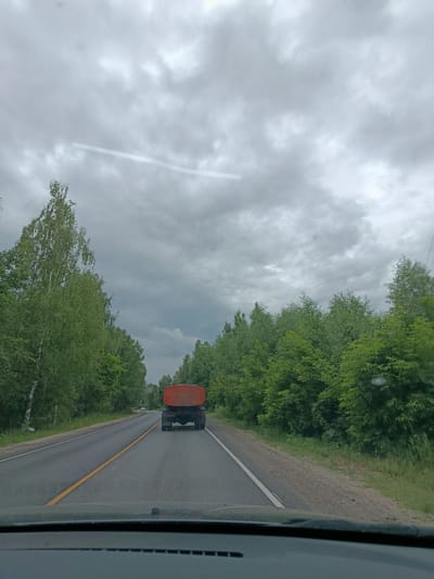 The image shows a view from inside a vehicle driving on a paved road. In the distance, a large orange tanker truck is traveling in the same direction. The road is flanked by dense green trees on both sides. The sky is overcast with gray clouds, suggesting a cloudy or possibly rainy day. A single white line and a yellow line mark the lanes of the road. The setting appears to be rural, given the abundant trees and the type of road. The overall mood is neutral, conveying a sense of travel on an ordinary day. There is no visible text in the image. The location context is provided as Тополиный, Russia.