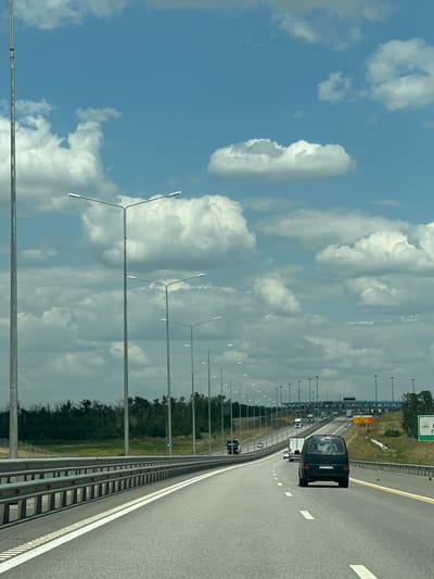 A wide, multi-lane highway stretches into the distance under a bright blue sky dotted with fluffy white clouds. The highway is lined with tall metal lamp posts on the left and a metal guardrail on the right. The road surface is asphalt, with clear white lane markings. Several vehicles are visible on the highway, including a dark colored van in the foreground, a white truck further ahead, and other cars and trucks in the distance. The landscape beyond the highway consists of green fields and a dense line of trees. In the far distance, there appears to be a toll plaza with multiple booths. The overall scene suggests travel on a sunny day in a suburban or rural area. The image is believed to be taken in the city of Bol'shoi Log, Russia, based on the provided context.