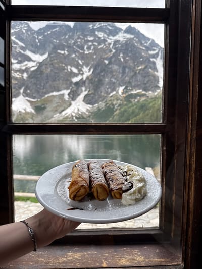 From an indoor vantage point in Brzegi, Poland, a white plate containing three cannoli, whipped cream, and powdered sugar is held in front of a window. The background shows the mountainous terrain of the surrounding area. The window is made of dark-brown wooden panes. The cannoli are topped with chocolate syrup. The hand holding the plate has a silver bracelet on its wrist.