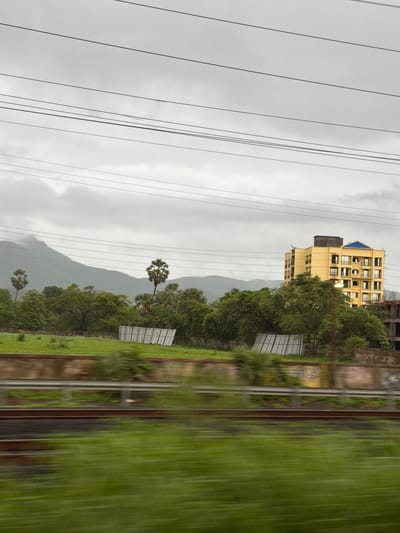 This image captures a view from a moving vehicle, likely a train, in Palghar, India, on a cloudy day. The foreground is blurred by motion, showing green vegetation and train tracks. In the mid-ground, a vibrant yellow, multi-story apartment building with balconies and a blue-tiled roof stands out amidst lush green trees. Behind the trees, a misty mountain range forms the backdrop under a heavily overcast sky. Several power lines crisscross the sky. There are also some white, panel-like structures in the mid-ground, possibly part of a construction or agricultural setup. The overall scene depicts an urban fringe environment with a blend of natural landscapes and developing infrastructure.