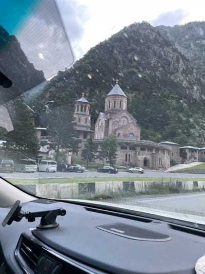 The image, captured from inside a vehicle, shows a view through the windshield of a stone church complex situated at the base of a steep, tree-covered mountain. The church features multiple domes, one prominent central dome topped with a cross, and a tall, separate bell tower also crowned with a cross. The architecture suggests an Orthodox Christian style. In front of the church, several cars are parked on a paved area. The foreground of the image is dominated by the dashboard of the vehicle, including a suction cup phone mount. The environment appears to be overcast with some visible water droplets on the windshield, suggesting rain or a wet surface. The location is identified as Dariali, Georgia, likely near the Dariali Gorge, known for its monasteries and natural beauty.
