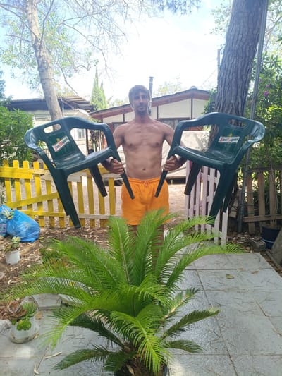 In Almardà, Spain, a shirtless man wearing orange swim trunks stands outdoors, holding two green plastic chairs, each bearing the "San Miguel" logo. He is positioned in front of a yellow picket fence, with a lush green plant in the foreground partially obscuring the view of his lower body. Behind him, structures with white walls and wooden accents are visible, along with trees and a clear sky. The ground is paved with grey tiles and scattered with debris. To the left of the man, a blue plastic bag lies on the ground.
