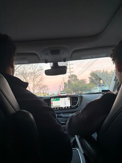 Inside a car, a passenger reclines in the back seat, head resting on the seat. The car's dashboard is visible with a GPS navigation display lit up. Outside, the sky is a soft blend of pink and gray during either a sunrise or sunset. Power lines run parallel to the road lined with trees. Another passenger is in the front seat, holding a phone towards the window.
