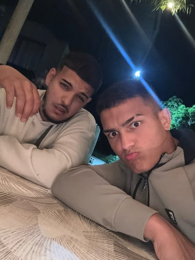 Two young men pose for a selfie outdoors at night. The man in the foreground wears a light-colored hooded sweatshirt, and rests his hand near his face. The man in the background wears a darker gray hooded sweatshirt. They are positioned in front of a surface with a radiating, sunburst-like pattern. The background shows glimpses of vegetation and a dark sky. The image was likely taken in Septemvritsi, Bulgaria.