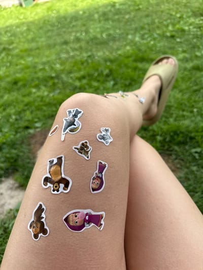 In Varzi-Omga, Russia, a person's leg, adorned with several cartoon stickers of bears and a girl, is visible. The stickers depict characters from the animated series "Masha and the Bear." The person is wearing a light-colored sandal on their foot. The background consists of green grass, suggesting an outdoor setting. The image is taken from a high-angle perspective, focusing on the leg and stickers.