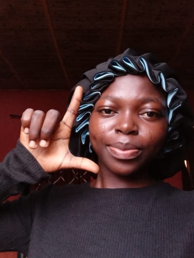A woman with dark skin and a black bonnet with light blue trim smiles slightly, looking directly at the camera. Her left hand is raised and angled above her head, forming a right angle with her fingers. She wears a long-sleeved, dark, ribbed shirt. The background is a mix of dark brown and red, with vertical patterns on the right side. The image is rotated, with the subject tilted to the left.