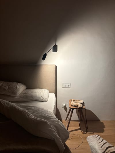 The image shows a bedroom in Celerina/Schlarigna, Switzerland. The bed is made with white linens and a brown headboard, with a white throw blanket draped over the top of the bed. A small wooden stool sits next to the bed, with a white fur throw on top. A wall-mounted lamp with a black cord is casting a light onto the bed. The wall is white with a light switch near the bed. The floor is made of wood. The environment is quiet and peaceful.  There are no people or other animals present in the image.