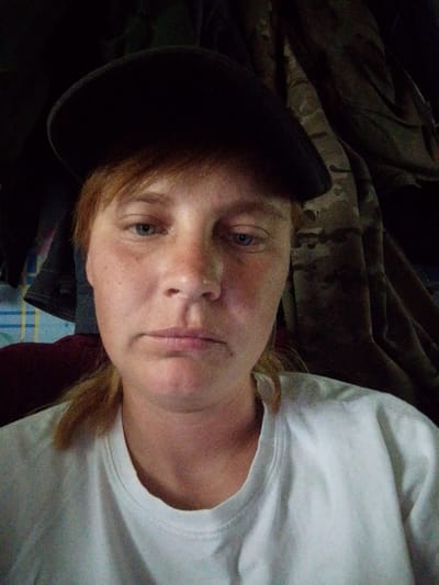 The image is a close-up, selfie-style photograph of a fair-skinned person wearing a dark baseball cap and a white t-shirt. The person has light-colored hair pulled back and visible around the ears. Their eyes are light blue, and they have a neutral or slightly downturned facial expression. The background is dark and appears to be fabric, with hints of camouflage patterns visible on the right side of the frame. The lighting suggests the photo was taken indoors, possibly in a dimly lit room. There are no identifiable landmarks or contextual clues to indicate the location in the city of Alekseevka, Russia.