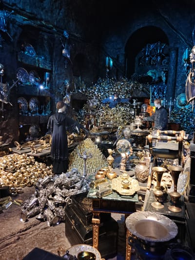 The image shows a room filled with an immense collection of various objects, predominately gold and silver, including goblets, plates, bars, orbs, and decorative pieces. Two mannequins are present, one wearing a dark hooded robe and another wearing a dark jacket and face mask. A man, also wearing a face mask, is partially visible amidst a pile of gold objects in the background. The room has stone walls with arched niches containing more displays of metal objects. The lighting is dim and theatrical, with a blue tint. The objects are arranged seemingly haphazardly across surfaces and on the floor. The location of this scene is likely at a Harry Potter exhibit in Three Rivers, United Kingdom.