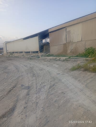 The image depicts an industrial or construction site in Bana Stream, Nigeria. In the foreground, a dirt road with prominent tire tracks is visible, suggesting recent vehicle activity. The road is uneven and appears to be composed of sand and small stones.
In the background, a long, corrugated metal building stands. The building has a light-colored, ribbed exterior. Part of the building's facade appears damaged or incomplete, with some sections missing or askew. To the right of the main structure, a section of the building is made of concrete blocks. Some greenery, including bushes and grass, is growing around the base of the building.
A piece of industrial machinery or equipment is partially visible inside an opening in the building. There are also pipes and wiring running along the exterior of the building. The sky above is clear with a hint of haze. The overall impression is of an operational or under-construction industrial facility. The timestamp "03/07/2026 17:42" is overlaid on the bottom right of the image. No people are visible in the scene.
