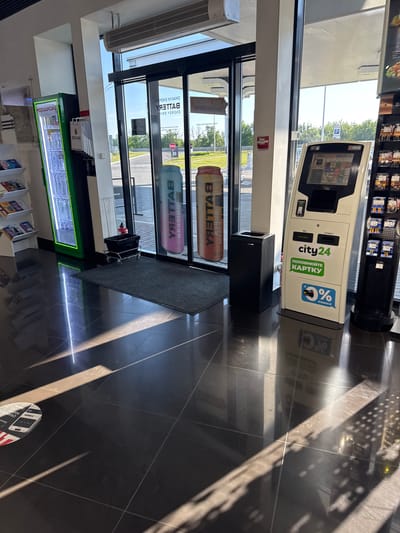 The image shows the interior of a convenience store, likely a gas station mini-mart, located in Dachne, Ukraine. The scene is brightly lit by natural light streaming through large glass doors and windows, creating strong reflections on the dark tiled floor.
In the foreground, the glossy black tiled floor dominates, with sunlight creating diagonal patterns of light and shadow. A circular sticker with red and white markings, possibly a floor marker or safety instruction, is visible in the lower left corner.
The middle ground features several key elements. To the left, a tall, illuminated green refrigerator case is filled with bottles. Beside it, a white display rack holds magazines or brochures. In the center, a black trash receptacle with wheels sits on a dark gray welcome mat in front of the glass doors. Behind the mat, two large beverage cans with "BATTERY" branding are visible, one blue and one a gradient of pink and orange.
To the right of the trash receptacle and mat is a modern, white self-service payment kiosk. The kiosk has a touchscreen interface with Ukrainian text and logos, including "city24" and "ПОПОВНЮЙТЕ КАРТКУ" (top up your card), and a "0% КОМІСІЇ" (0% commission) sticker. A black trash bin with a rectangular opening is placed next to the kiosk. Further to the right, a black display unit with multiple shelves is stocked with packaged goods, appearing to be tobacco products or small food items.
The background shows the exterior through the glass doors, revealing a paved area, greenery, and a blue "P" parking sign against a clear sky. The overall impression is one of a clean, well-lit, and modern retail environment. There are no people visible in the image.