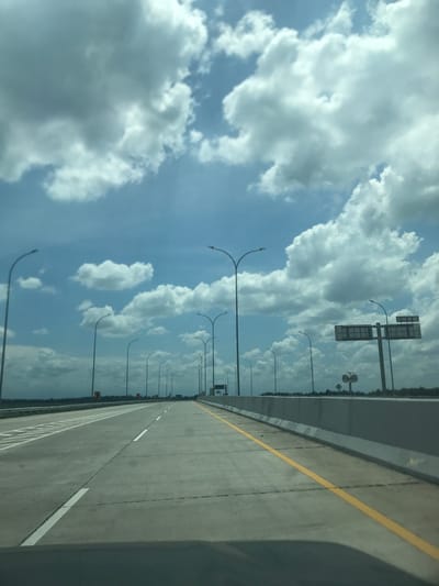 The image shows a highway with a blue sky and clouds on the left. Street lights line the side of the road. The highway has multiple lanes, marked with yellow and white lines. There are overhead signs and barriers on the right side of the road. The exact location in Stabat, Indonesia is not discernible from the image.