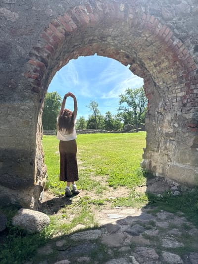 A woman with long, brown hair stands in a field, facing away from the camera. She is wearing a white t-shirt and a brown and black leopard-print skirt, along with grey and white socks and dark shoes. Her arms are raised above her head, hands clasped, as if stretching. She is positioned within an arched stone and brick ruin. The background features a bright blue sky with wispy white clouds, lush green grass, and trees in the distance. The scene appears to be set during the daytime under fair weather. The stone and brick structure suggests a historical or ancient location. The overall mood is peaceful and serene. No visible text is present in the image. The location context is Dobele, Latvia.