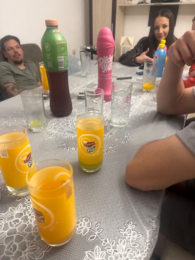 The image shows a gathering of people around a table covered with a gray tablecloth with a white floral pattern. The table holds several glasses and bottles, including a green and brown bottle, two clear glasses, a pink bottle, and three yellow Lipton Ice Tea glasses, filled with a yellow liquid. Three individuals are visible: a woman pouring a blue bottle of liquid into a glass, a man in a green shirt is visible in the bottom left corner, and the arm and shoulder of another person are in the upper right corner.
The setting appears to be indoors. There is no verifiable evidence to confirm this location is Graf Ignatievo, Bulgaria.