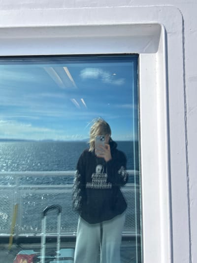 A vertical, eye-level shot captures a mirror selfie taken by an individual reflected in a large window aboard a vessel, likely a ferry, navigating open waters near Point Roberts, United States. The subject, a person with blonde hair, is visible from the upper chest to mid-thigh, their face obscured by a silver-colored smartphone held to capture the reflection. They wear a black long-sleeved hoodie with white graphics and text on the front and sleeves, paired with light-colored, wide-leg pants.

The reflection reveals the exterior environment: a vast expanse of deep blue, rippling ocean under a clear blue sky with scattered white clouds. Distant landmasses, appearing as dark, low-lying hills or mountains, are visible on the horizon. A white metal railing, consistent with a boat's deck, is reflected across the lower mid-ground. In the lower-left portion of the reflection, a silver-colored rolling suitcase with a black handle and a blue soft-sided bag are discernible. The interior elements reflected include white structural framing around the window and elongated white reflections on the glass, possibly from overhead lighting or panels within the vessel. The sunlight suggests a bright day with visible wind effects on the person's hair.