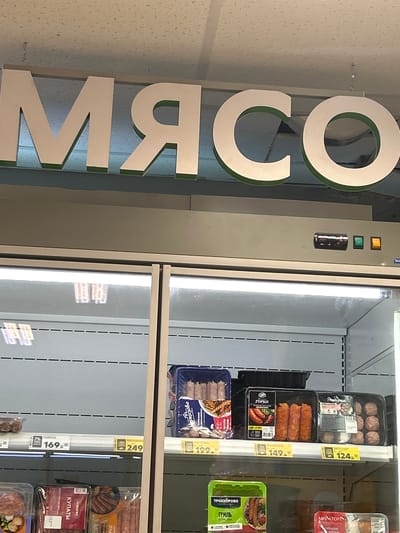 A sign with large white letters spelling "МЯСО" (meat in Russian) is visible above a refrigerated display case in a store. The display case contains various meat products, including sausages and pre-packaged meals. Prices are displayed on yellow tags beneath the products. The environment appears to be a supermarket in Lotoshino, Russia.