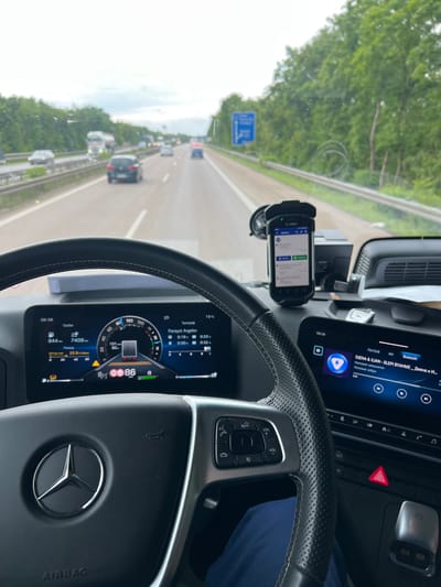 The image depicts the interior of a Mercedes vehicle, likely a truck, traveling on a highway. The driver is in transit. The dashboard displays information, including a speedometer reading 86 km/h, fuel consumption, and a tachograph. A mobile device mounted on the dashboard shows a screen. The vehicle's infotainment system is playing music. The scene indicates the vehicle is on a highway with other cars, possibly in Germany, as the dashboard display includes "Panayot Angelov".