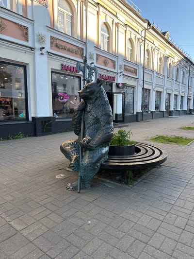 The image shows a bronze statue of a bear sitting on a sidewalk in front of a building. The bear is holding a staff with an axe head. It is a sunny day, and the light is casting shadows on the ground. The building in the background has a light-colored facade with arched windows and ornate decorations. There are storefronts with signage on the ground floor. A round bench with a planter in the center is located next to the statue. The sidewalk is paved with rectangular bricks. The scene is set in the city of Feklino, Russia.