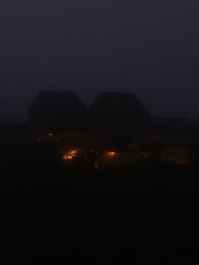 The image shows a dark, foggy scene with two dimly lit buildings in the foreground. The buildings appear to be traditional, with steep roofs. There are visible lights within the buildings, suggesting occupancy. The surrounding environment is obscured by darkness and fog, making it difficult to discern specific details of the location. The overall impression is one of a quiet, rural setting at night. While the image is labeled as being in Marhi, India, there are no specific visual cues in the photograph to confirm this location.