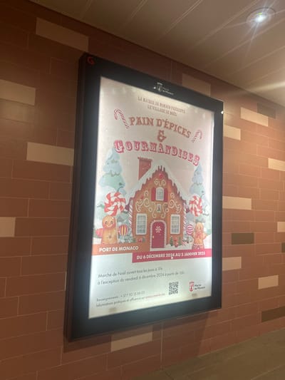 An illuminated sign lies flat on a tiled floor in what appears to be an indoor public space in Beausoleil, France. The floor consists of light brown and beige rectangular tiles arranged in a linear pattern. A bright overhead light fixture is visible on a paneled wall to the left, casting a glow.
The sign is a promotional poster for a Christmas event, featuring a festive illustration of a gingerbread house adorned with white frosting, candy canes, gingerbread figures, and other sweets against a light blue and white background.
Visible text on the sign indicates:
- "LA MAIRIE DE MONACO PRÉSENTE LE VILLAGE DE NOËL" (The Monaco City Hall Presents The Christmas Village)
- The main title reads "PAIN D'ÉPICES & GOURMANDISES" (Gingerbread & Delicacies).
- The location is specified as "PORT DE MONACO."
- The event dates are "DU 6 DÉCEMBRE 2024 AU 5 JANVIER 2025" (From December 6, 2024 to January 5, 2025).
- Details about the "Marché de Noël" (Christmas Market) state it is "ouvert tous les jours à 11h à l'exception du vendredi 6 décembre 2024 à partir de 16h" (open every day at 11 am, with the exception of Friday, December 6, 2024, starting at 4 pm).
- Contact information includes "Renseignements: +377 93 15 06 03" and a website for "Informations pratiques et affluence sur www.mairie.mc."
- The "Mairie de Monaco" logo is present in the top left and next to a QR code on the right.
The sign is brightly lit from within, contrasting with the slightly dimmer surroundings. No people are visible in the image.