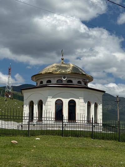 A white, octagonal building with a golden dome and a crescent moon finial stands in the foreground. The building has arched windows and is surrounded by a black metal fence. Behind the fence, a green grassy hill slopes upwards. In the background, mountains covered in trees rise against a blue sky with white clouds. A tall red and white lattice tower, likely a communication tower, is visible on the left side of the frame. The scene is set in a rural or mountainous area, possibly on the outskirts of the city of Verkhnyaya Teberda, Russia.