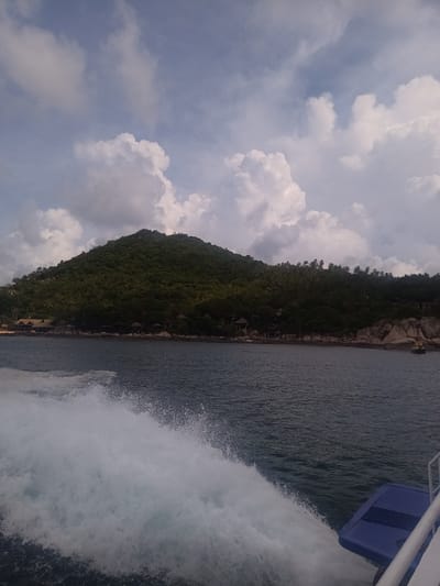 The image shows a view from a boat approaching a lush, green island. The sky is partly cloudy with large, white cumulus clouds. The island is covered in dense vegetation, possibly a tropical forest. At the base of the island, along the coastline, there are some visible structures that appear to be buildings or houses nestled among the trees and rocks. The foreground of the image is dominated by the choppy, dark blue water of the sea. White foam from the boat's wake is visible in the water. A portion of the boat itself is visible in the lower right corner, revealing a blue platform. Given the mention of Mae Haad, Thailand, it is reasonable to assume that the island is located in the vicinity of Mae Haad.