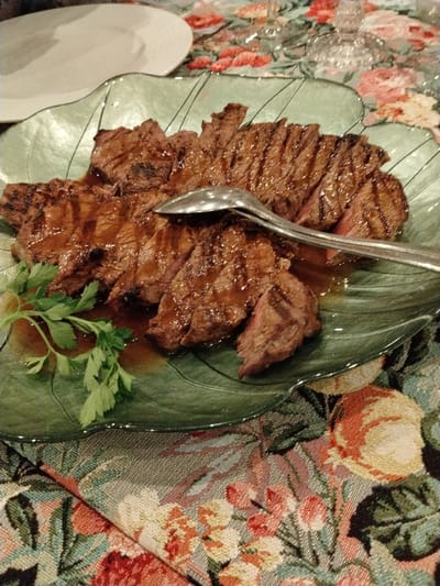 Sliced, grilled steak covered in sauce sits on a light green, leaf-shaped platter. A serving spoon rests on the meat. A sprig of parsley adds a touch of green to the dish. The platter is placed on a table covered with a floral tablecloth. Another plate, presumably also part of the meal, is visible in the upper-left corner. The overall setting suggests a formal or special occasion.