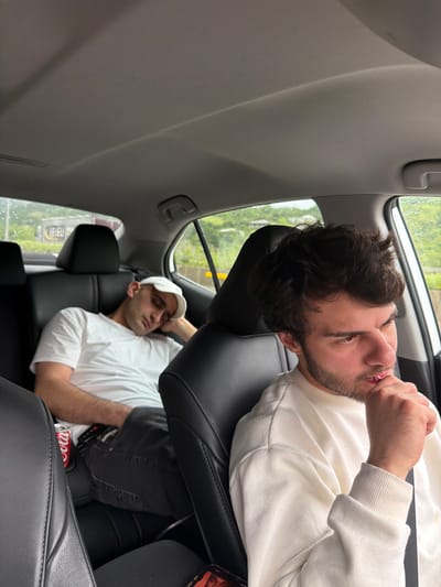 Two men are in a car. The man in the foreground is facing the camera and appears to be eating something. He is wearing a white sweatshirt. The man in the background is wearing a white t-shirt and a white baseball cap. He appears to be sleeping. The car window is visible and it is raining outside. The location is likely Grigalati, Georgia.
