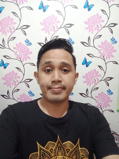 A man with dark hair, a mustache, and light skin faces the camera. He wears a black t-shirt with a golden design on the front. The background is a patterned wallpaper with pink flowers, gray leaves, and blue butterflies. The man's expression is neutral. The light appears to be evenly distributed across the scene.