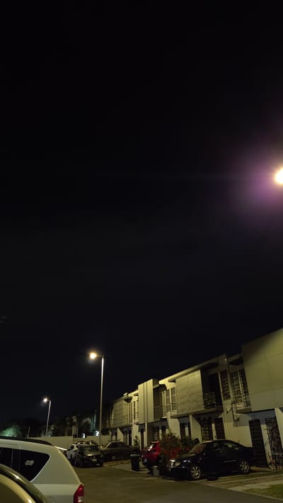 This image captures a residential street at night within Fraccionamiento Real del Valle, Mexico. The scene is illuminated by multiple artificial streetlights under a dark, clear sky. A row of two-story, modern townhouses with light-colored exteriors and balconies lines the right side of the paved street. Numerous vehicles are parked on both sides of the street and in front of the residences. In the immediate left foreground, the rear section of a white SUV is visible. Further down, on the left, a dark-colored car is parked. On the right, directly in front of the townhouses, a dark sedan, a red SUV, and another dark sedan are parked. Two tall streetlights, emitting bright white-yellow light, are prominent fixtures, with one in the mid-ground and another further down the street on the left. Sparse vegetation, including bushes and small trees, is present along the property lines. A dark waste bin and a red child's bicycle are visible on the right side, near the houses. No individuals are present in the frame.