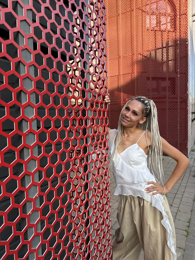 A woman with blonde braided hair is standing next to a red hexagonal metal fence. She is wearing a white ruffled top and loose beige pants. The background shows a brick wall and a building with red perforated metal siding. The scene appears to be outdoors in an urban or semi-urban environment. The location is stated to be Есбол ауылдық округі, Kazakhstan.