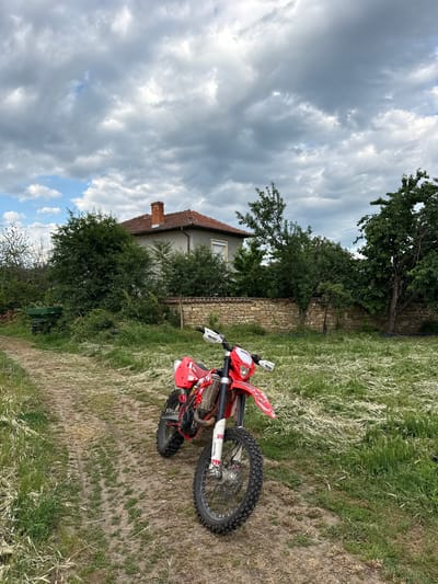 An outdoor scene in Bratya Daskalovi, Bulgaria, features a red and white dirt bike (motorcycle) parked in a grassy, unpaved area. The image is oriented sideways, with the ground appearing on the right and the sky on the left.

The main subject is the dirt bike, prominently red with white accents, including "Beta" branding visible on the handguards and front fender. Its knobby tires suggest off-road use. Letters like "SK" are visible on the front fork. The bike is resting on its kickstand on a dirt path next to an area of tall, dry and green grass and weeds.

In the background, a traditional-looking house with a tiled roof and a brick chimney is partially visible, nestled among green trees. A stone wall runs along part of the property line, separating the house area from the grassy field where the bike is located. A green bench is partially visible in the distance near the house.

The sky is largely overcast with significant gray clouds, but patches of blue sky are also visible, suggesting a break in the weather or an intermittent cloud cover. No people are present in the image. The setting appears rural or semi-rural, typical of a countryside environment. The time of day is daytime, likely late morning or afternoon given the light.