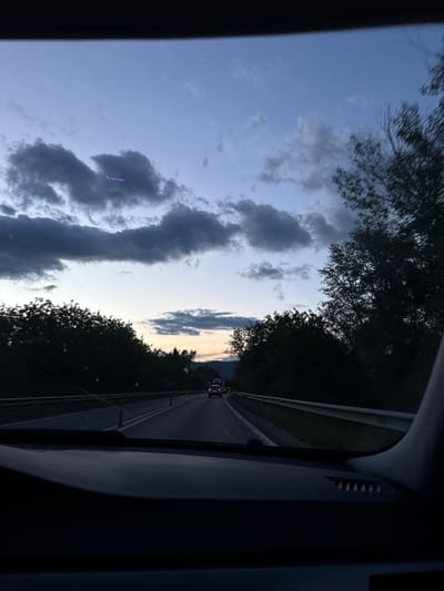 The image depicts a view from inside a vehicle, likely a car, driving on a road. The sky is visible through the windshield and side window, showing a sky with scattered clouds. Trees line both sides of the road. In the distance, other vehicles are visible on the road ahead. The interior of the vehicle is dark, with parts of the dashboard and window frame visible. The location is unknown. There is no evidence that the location is in the city of Poleto, Bulgaria.