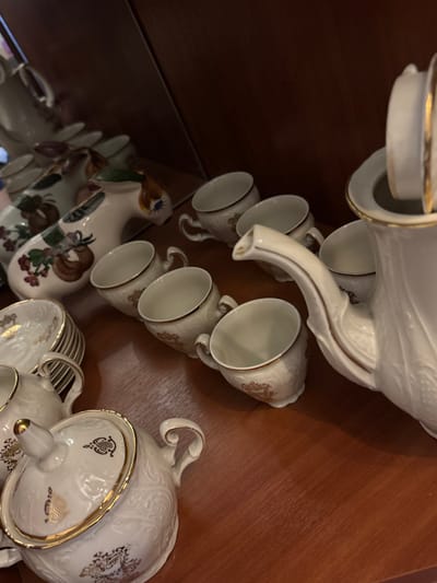 The scene is located in Velikiye Luki, Russia. It depicts a collection of ceramic tableware arranged on a polished, reddish-brown wooden surface within an indoor setting.
On the right side of the frame, a white porcelain coffeepot or teapot with a gold-trimmed lid and spout is prominently featured. To its left and in front, six matching small cups, also white, are visible, each adorned with a gold rim and intricate embossed or painted patterns. In the lower left quadrant, a lidded sugar bowl and a smaller creamer, both white with gold details and decorative designs, are present, along with a stack of matching saucers.
Towards the upper left background, at least two distinct, elongated, animal-shaped ceramic pitchers are arranged. These vessels are white with hand-painted floral motifs and exhibit brown and green accents, including a visible painted eye on one.
No individuals are present, and no actions or interactions are depicted; the objects are static under ambient indoor illumination, casting soft shadows on the wooden surface.