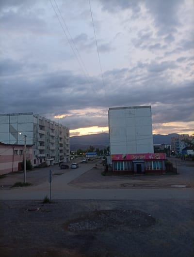 The image shows a street scene in the city of Shagonar, Russia, at dusk. The sky is overcast with dark, heavy clouds, but a sliver of orange and yellow light from the setting sun is visible near the horizon behind a range of low hills. In the foreground, a paved road runs horizontally across the frame, with a gravelly area to the left and right.
To the left, a light pink and beige multi-story residential building is partially visible. To its right, and dominating the mid-ground, is a tall, rectangular, light gray apartment building with multiple floors and balconies. In front of this building, there is a low, rectangular structure with a red trim and large windows, which appears to be a shop or business. It has colorful signage on its facade, predominantly pink and yellow.
Several cars are parked or driving on the road in the mid-ground, including two dark-colored sedans and a lighter-colored vehicle. A street sign on a pole is visible in the middle of the road. Further in the background, other residential buildings and some sparse vegetation can be seen. The overall impression is of a somewhat bleak, utilitarian urban environment under a dramatic sky.