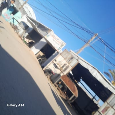 The image shows a street in Al-Mawasi, Unknown, with a series of damaged or dilapidated structures lining the road. The structures appear to be shops or stalls, with metal roofing and open fronts. There is at least one person visible in the background. Power lines crisscross the sky above. The street is paved and appears to be in a state of disrepair.