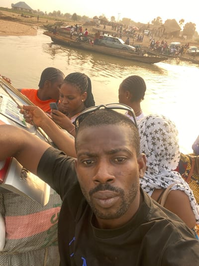 A man takes a selfie in what appears to be a boat or ferry, with other passengers visible in the background. A crowd of people and vehicles, including a car, are visible on land in the distance. The man wears a black shirt and has sunglasses on his head. Two women stand beside him looking at a license plate. The surroundings suggest a riverside or lake setting, with a dusty or sandy shoreline visible on the horizon.