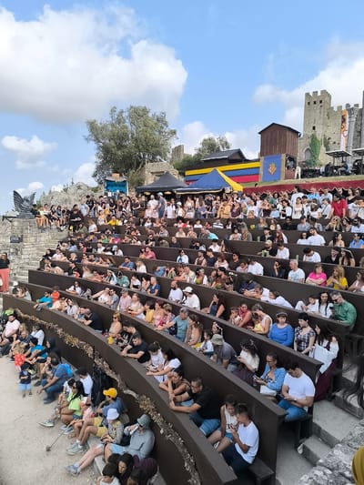 The image captures an outdoor audience seated in tiered wooden bleachers, facing a performance area. The setting appears to be within the medieval walls of Óbidos, Portugal, suggested by the presence of ancient stone structures and a castle in the background.

In the foreground, the bleachers are filled with a diverse crowd of people of various ages, all facing in the same direction, indicating they are watching an event. Many individuals are holding smartphones, some appearing to be engaged with them, while others are looking towards the unseen performance. The seating is organized in concentric semi-circles, with each row slightly elevated from the one in front. The bleachers themselves are constructed from dark wood and are decorated with what looks like dried foliage or garlands.

Behind the audience, the landscape transitions to rocky terrain and vegetation. Further back, several buildings and structures are visible. A tall, stone castle with battlements is prominently featured on a hill, illuminated by the sunlight. Adjacent to the castle are wooden structures and tents, one of which displays a colorful banner with horizontal stripes of blue, yellow, and red, and a coat of arms. Another banner with a dragon motif is also visible. The sky is a bright blue with scattered white clouds, suggesting a clear, sunny day.

In the upper left quadrant of the image, a dark, stylized sculpture of a winged creature, possibly a dragon or griffin, is mounted on a stone platform. The overall impression is of a cultural or historical reenactment event taking place in a historically significant location.