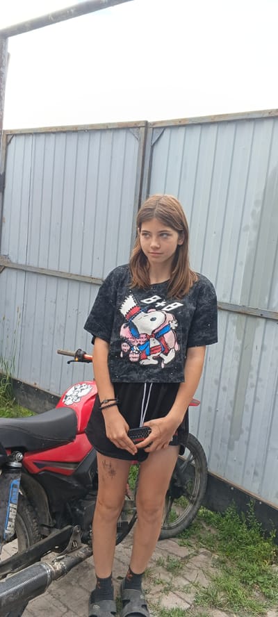 In Chernyavskii, Russia, a young woman stands in front of a corrugated metal fence. She is wearing a black crop top with a graphic print of Snoopy, and black shorts with white drawstrings. She has on dark socks and black sandals. On her left wrist, she wears two beaded bracelets. She is holding a small, dark object, possibly a phone case, in her hands. To her left, a red motorcycle is parked. The ground is a mix of brick pavers and grass. The overall environment suggests an outdoor, possibly rural or suburban, setting.