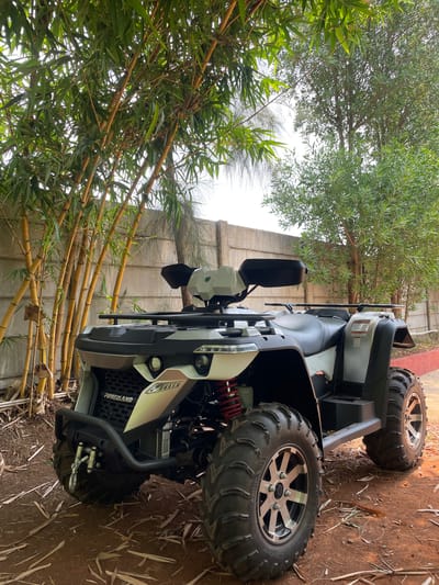 An ATV (All-Terrain Vehicle) or quad bike, predominantly white and black, is parked outdoors on reddish-brown dirt ground. The vehicle features large, rugged off-road tires with silver multi-spoke rims, prominent red suspension components, black side mirrors, and black utility racks at the front and rear. The brand "POWERLAND" is clearly visible on the ATV's front grille, and "GEAR LITE" is discernible on the tire sidewalls.
The setting is an outdoor environment with lush vegetation. On the left side of the frame, a dense cluster of vibrant green bamboo plants with golden-brown stalks extends upwards, partially obscuring a light gray concrete wall in the background. In the upper part of the image, a tree with smaller, dark green leaves is visible. The ground surrounding the ATV is uneven, covered with sparse dry leaves and plant debris.
The bright, diffused lighting suggests it is daytime. The scene evokes a natural or semi-rural outdoor setting, consistent with the Dhundshi, India location. No individuals are present, and the ATV appears stationary, implying it is either parked or awaiting use.
