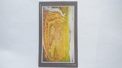 The image is a vertical photograph of a horizontally oriented, framed painting, displayed against a plain white background. The painting, contextually associated with the city of Limpok, Indonesia, depicts a stylized urban landscape. On the right side of the artwork, large expanses of bright yellow and green suggest open land or fields. Towards the center, a dense, sprawling collection of abstract orange and brown structures represents a city or settlement, with numerous buildings appearing to rise from the landscape. On the left, a prominent winding body of water, rendered in shades of blue and white, resembling a river or canal, flows through or alongside the urban area. The overall artistic style is impressionistic or abstract, focusing on color and form rather than fine detail. No discernible individuals, specific actions, or interactions are visible within the artwork.