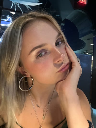 A young Caucasian female, with long blonde hair parted slightly to her left, occupies the majority of this close-up portrait. She has a fair complexion with visible freckles across her nose and cheeks, green eyes, and is wearing pink blush, purple eyeshadow, and lip gloss. Her right hand is raised to support her chin, fingers gently resting against her jawline. She wears a dark-colored, possibly green or black, spaghetti strap top. Her visible jewelry includes a large silver hoop earring on her left ear, multiple layered silver necklaces—one with small beads and another with a green teardrop-shaped pendant—a silver ring with an intricate design on her right ring finger, and a dark blue beaded bracelet on her left wrist. The background is dimly lit, featuring elements of modern interior design. Above her head, a white, abstract, wave-like ceiling fixture is visible, alongside blue accent lighting and a partially visible blue neon sign. In the upper right corner, an illuminated red "EXIT" sign with white lettering is prominent. This scene is identified as taking place inside Finn's Restaurant, located on Wharf Street in Victoria, British Columbia, Canada.