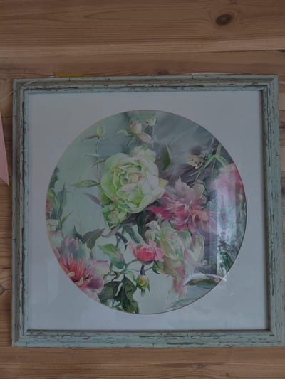 The image shows a framed circular watercolor painting of peonies, displayed on a wooden wall. The painting features a variety of peony blossoms in shades of light green, pink, and white, with dark green leaves and some buds. The frame is light blue-green with a distressed, chippy paint finish. The wooden wall behind the painting has visible grain and knots. The scene appears to be indoors. There is no discernible location in the city of Бярэзінскі сельскі Савет, Belarus.