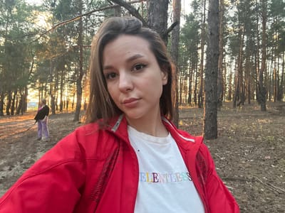 The image is a selfie taken outdoors in a wooded area, likely a forest, with tall trees. The photo is oriented sideways. In the foreground, a person is looking directly at the camera. They are wearing a white t-shirt with multi-colored text and a red jacket. The text on the t-shirt appears to read "RELENTLESS" in colorful, embroidered lettering. The person's facial expression is neutral. In the background, another person, possibly a child, is visible wearing purple pants and a dark top. They are further back in the scene, appearing smaller in scale. The setting suggests it is late afternoon or early evening due to the warm, golden light filtering through the trees, casting long shadows and creating a sunset or sunrise effect. The ground is covered with dry leaves and some patches of grass, indicating it might be autumn or a dry season. The overall mood of the image is casual and relaxed, capturing a moment in nature.