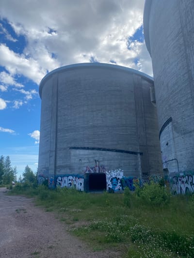 The image shows a view of the concrete silos at the former Sunila Pulp Mill in Kotka, Finland. The silos are cylindrical structures with visible vertical joints in the concrete. The larger silo in the foreground is covered in graffiti, including the word "SASHO" in large white letters. There is an open entrance to the silo at ground level, also surrounded by graffiti. The ground in front of the silos is a dirt path with patchy green grass and small white flowers growing in it. To the left of the silos, there are trees and a vehicle is partially visible in the distance. The sky is blue with scattered white clouds. The image is taken from a low angle, emphasizing the scale of the silos.