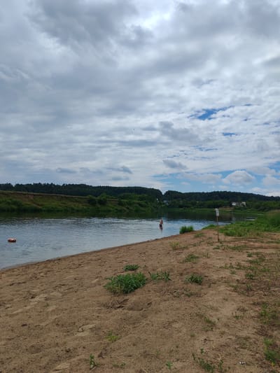 A wide shot captures a serene riverside scene under a cloudy sky. In the foreground, a sandy bank slopes gently towards the calm, blue water. Scattered on the sand are small green plants, indicating a natural, undeveloped area. To the left of the frame, a small, orange buoy floats in the water near the bank. Further out in the river, a lone figure, appearing to be a child, stands in the water. The opposite bank of the river is lined with a dense, green forest, with some houses visible in the distance through the trees. The sky is filled with a mix of gray and white clouds, with patches of blue sky peeking through. A wooden post with a sign is visible on the right side of the sandy bank. The scene depicts a natural recreational spot, likely a public beach or swimming area, in the countryside of Палыкавіцкі сельскі Савет, Belarus.