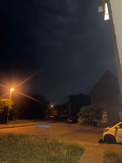 A night-time outdoor shot captures a residential street scene in the Borough of Swale, United Kingdom. A prominent street lamp illuminates the foreground with a warm glow, casting long shadows and highlighting a patch of tall, dry grass along the edge of the pavement. To the right, the brick facade of a house is visible, with a white car parked on the right side of the road, its front wheel partially on the grass verge. Further down the street, a road sign reads "MULBERRY WAY." Several cars are parked or driving on the road, and glimpses of houses and trees suggest a suburban environment. The sky above is dark, with visible clouds.